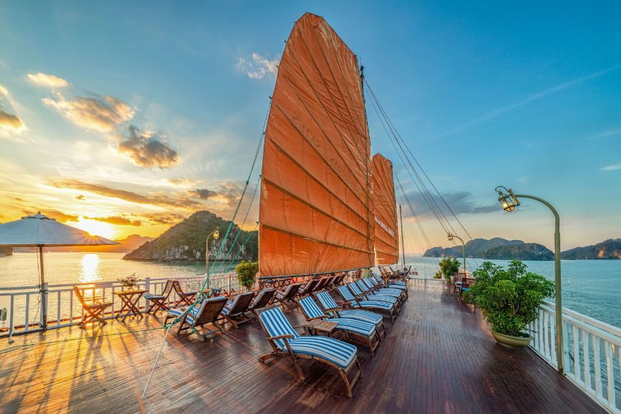 Wooden sundeck at sunset with orange sails, blue-striped sun loungers, and karst mountain views
