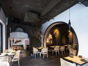 Modern dining space with black arched doorways, white rattan chairs, exposed ceiling, and tree decor