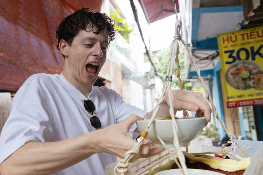 Tourist eating street-side hu tieu noodles from a hanging bowl stall