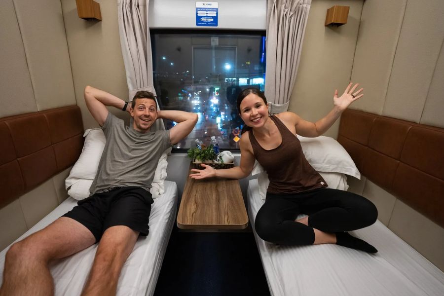 Couple relaxing in a private VIP tourist cabin on an overnight train in Vietnam with city lights outside the window